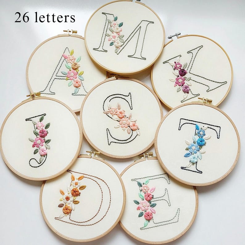 Op de afbeelding: Een verzameling geborduurde letters, elk in een houten hoepel. De letters zijn omrand met zwarte draad en versierd met kleurrijke bloemenborduurwerken. De tekst "26 letters" is zichtbaar bovenaan.