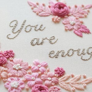 May include: Embroidered artwork with the words "You are enough" in gray thread. The design features pink and light pink flowers and leaves, creating a floral border and accents. The background is a light cream color.