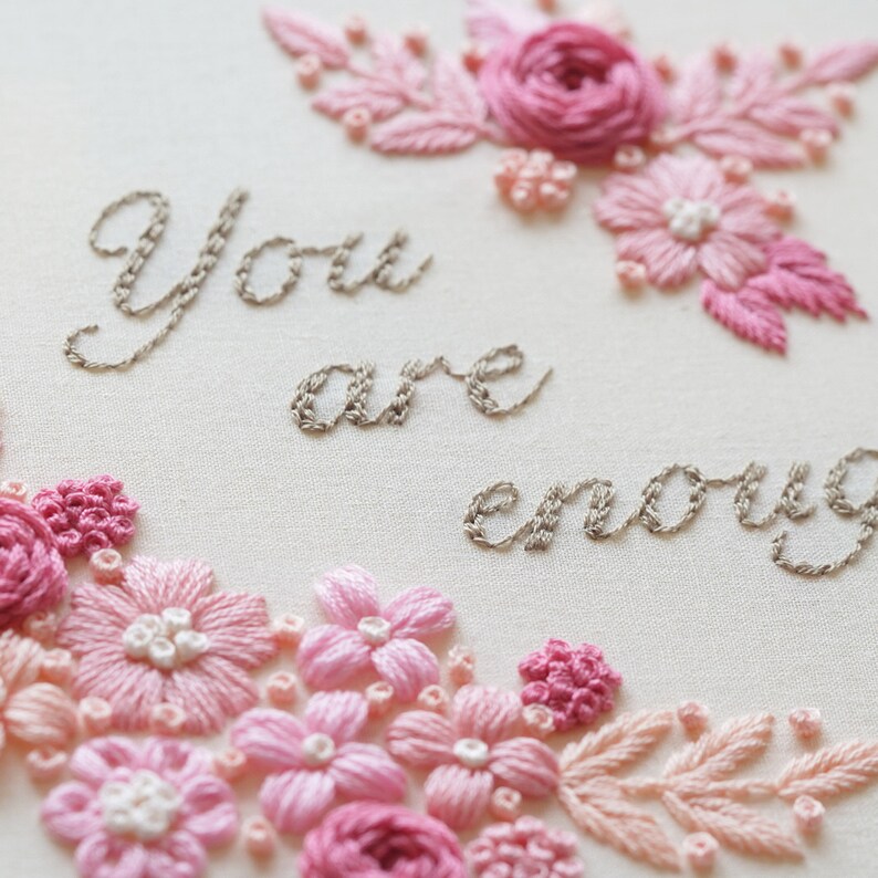 May include: Embroidered artwork with the words "You are enough" in gray thread. The design features pink and light pink flowers and leaves, creating a floral border and accents. The background is a light cream color.