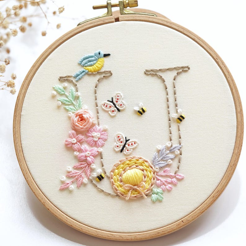 May include: Embroidered hoop art showing the letter "U" with a bird, butterflies, bees, and flowers. The design includes a blue and yellow bird, pink and peach flowers, and a yellow nest. The embroidery is set in a wooden hoop.