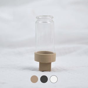 Könnte beinhalten: Ein klares Glasfläschchen mit beigefarbenem Sockel. Das Fläschchen ist zylindrisch mit abgerundeter Oberseite. Der Sockel hat eine solide, rechteckige Form mit einem kreisförmigen Ring, der das Fläschchen hält. Der Hintergrund ist weiß.