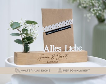 May include: A wooden block with the words "Alles Liebe" and the names "Janina & Bennet" engraved on it, along with the date 24.09.2026. A small vase with greenery and a card with a decorative ribbon are also present.