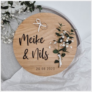 Könnte beinhalten: Eine Holz-Ringkissenbox mit den Namen "Meike & Nils" und dem Datum "26.08.2023" eingraviert. Zwei goldene Ringe sind mit einem weißen Band zusammengebunden und auf der Box platziert. Ein kleiner Strauß weißer Blumen liegt auf der Box.