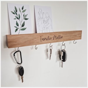 Könnte beinhalten: Ein Schlüsselbrett aus Holz mit der Gravur "Familie Müller". Das Brett hat sechs Haken und ist an einer weißen Wand montiert. Über dem Brett hängen zwei gerahmte Bilder, eines mit einer grünen Pflanze und das andere mit einer Strichzeichnung einer Familie.
