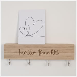 Könnte beinhalten: Ein Schlüsselbrett aus Holz mit fünf Haken und dem eingravierten Text "Familie Benedikt". Auf dem Schlüsselbrett liegt eine weiße Karte mit einer Zeichnung von zwei ineinander verschlungenen Herzen.