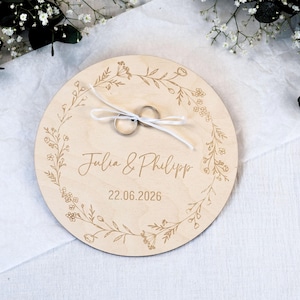 Könnte beinhalten: Ein runder, hellfarbiger Holzringhalter mit floralem Kranzdesign. Die Namen "Julia & Philipp" und das Datum "22.06.2026" sind eingraviert. Zwei Eheringe sind mit einem weißen Band befestigt.
