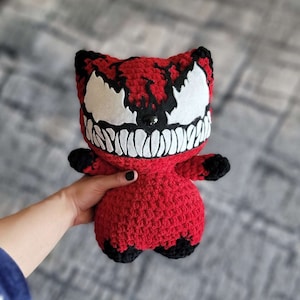 Puede incluir: Un juguete de peluche rojo de crochet con forma de gato con una cara inspirada en Venom en blanco y negro. El juguete tiene ojos negros y una amplia sonrisa blanca.