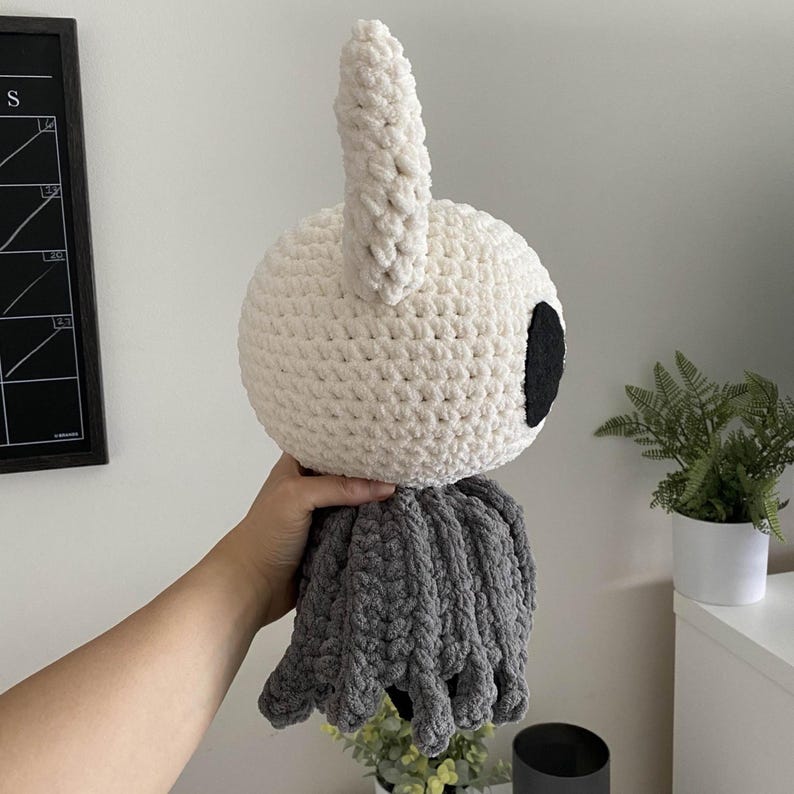 May include: A large, handmade crochet plush toy in the shape of a character with a white head, a black eye, and a gray lower body. The plush has a large, upright ear. The toy is held up against a white wall.