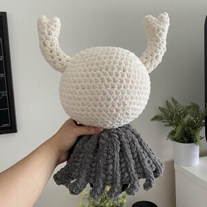 May include: A handmade crochet crab toy with a white head and two curved, upward-pointing claws. The crab's body is a fluffy, round shape, and the lower portion is a gray, fringed skirt. The toy is soft and plush.