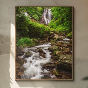 May include: Framed photograph of a waterfall cascading down a rocky cliff, surrounded by lush green trees and foliage. The water flows into a stream with large rocks. The image is in a wooden frame.