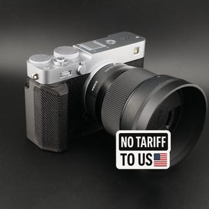 May include: A silver and black digital camera with a large lens and a black lens hood. The camera has several dials and buttons. A sticker on the lens hood reads "NO TARIFF TO US" with a small American flag.