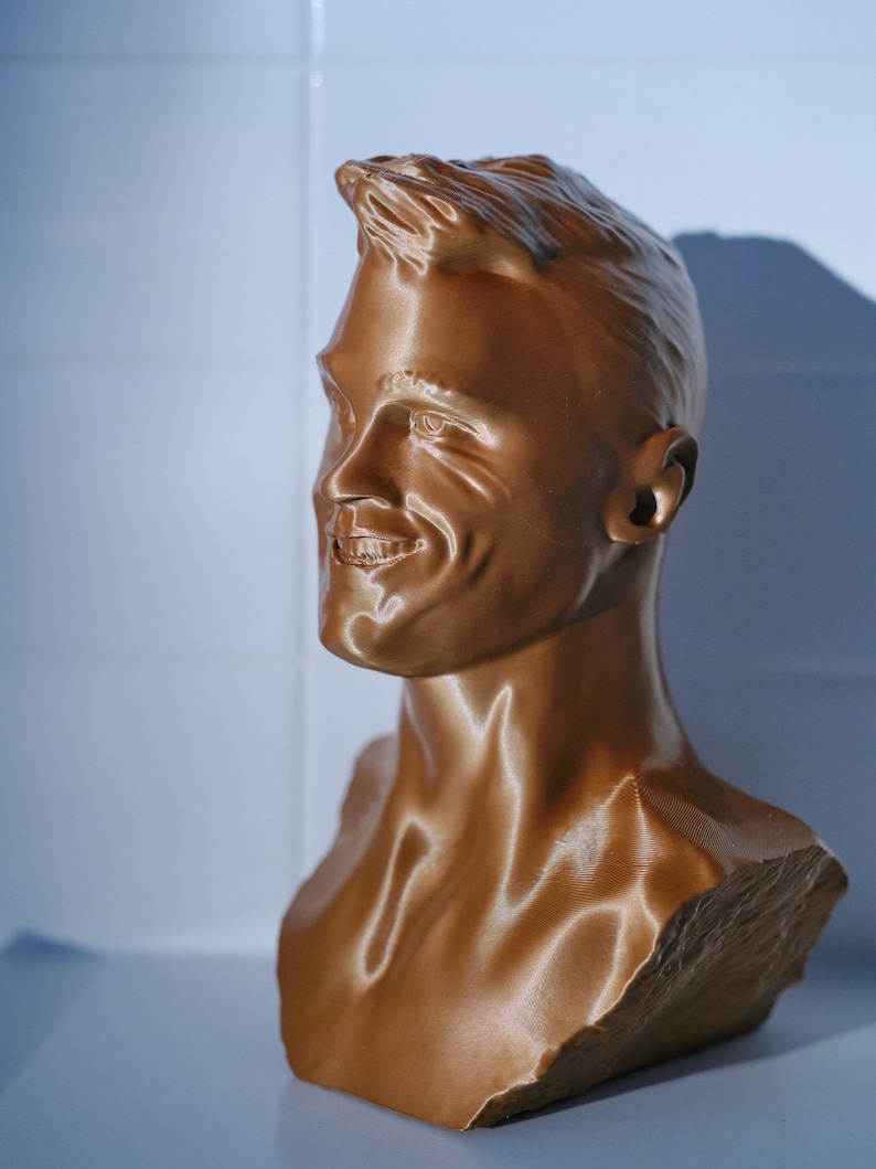 May include: A bronze-colored bust of a smiling person. The sculpture features detailed facial features, including hair, eyes, nose, and mouth. The bust is a three-dimensional representation of a person's head and upper chest.