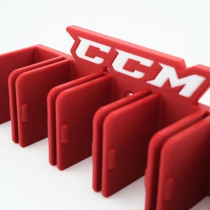 Puede incluir: Porta palos de hockey de pl&aacute;stico rojo con seis ranuras y el logotipo de CCM en blanco.