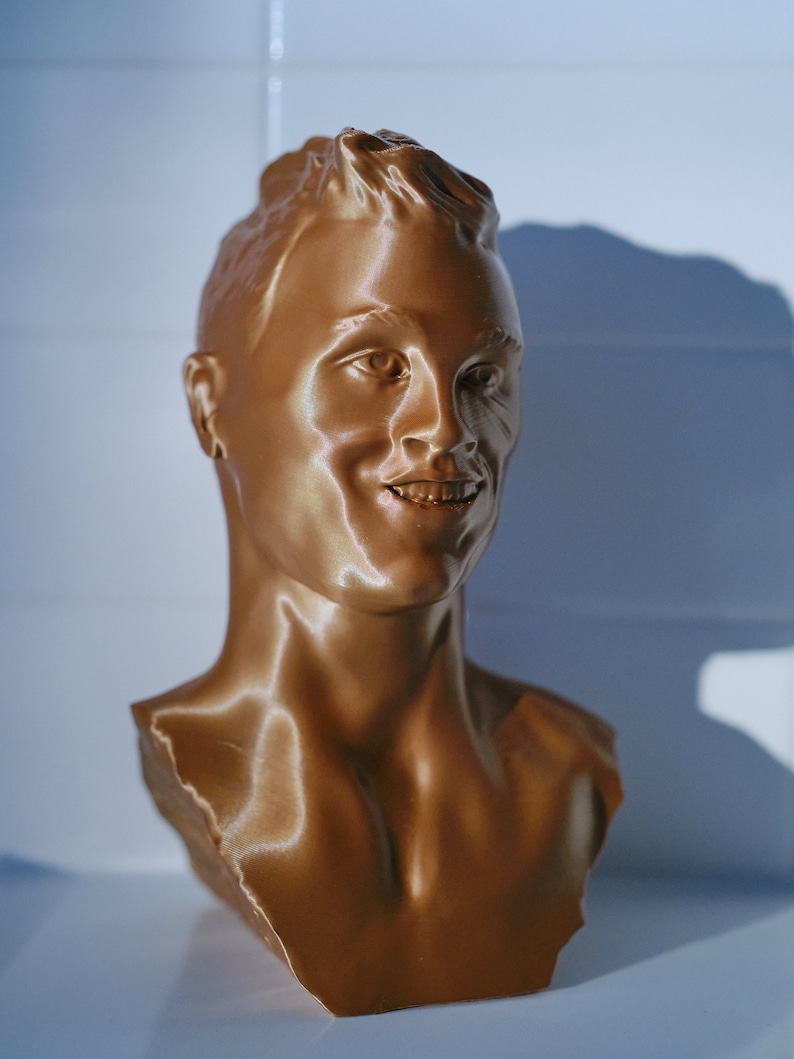 May include: A bronze-colored bust of a person with short hair and a smiling expression. The sculpture is a detailed representation of a human face and upper torso, with visible facial features and neck.