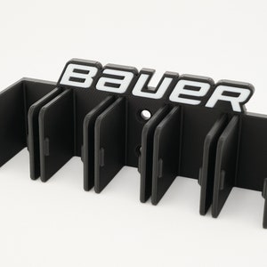Puede incluir: Porta palos de hockey de pl&aacute;stico negro con seis ranuras y la palabra "BAUER" en letras blancas.