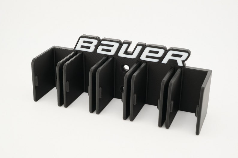 Puede incluir: Porta palos de hockey de pl&aacute;stico negro con seis ranuras y la palabra "BAUER" en letras blancas.
