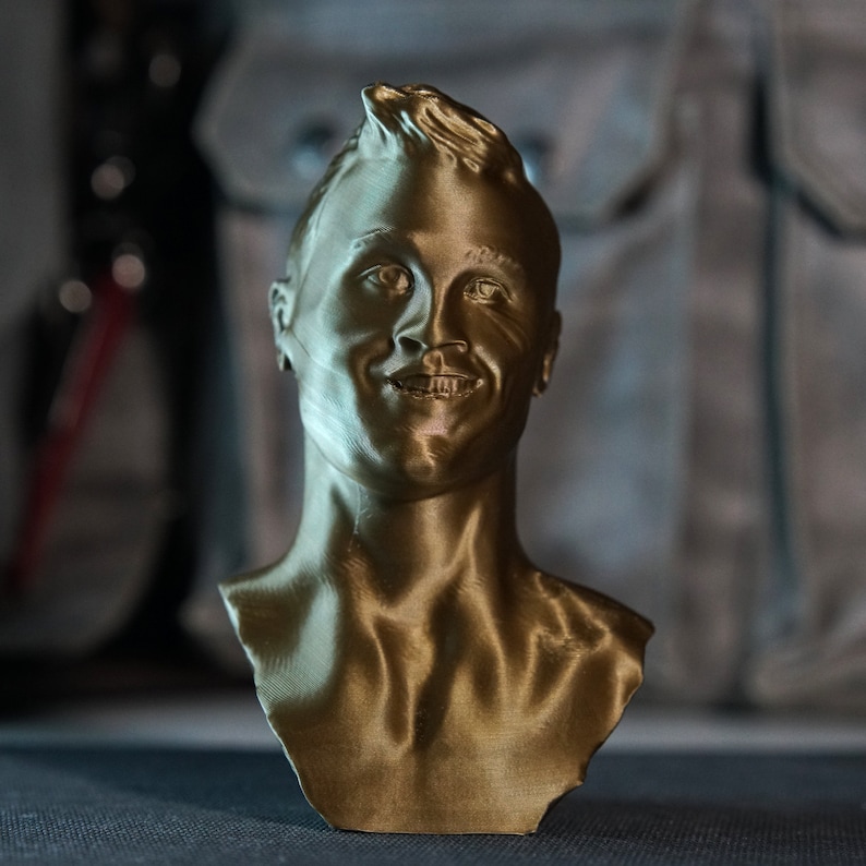 May include: A gold-colored bust of a smiling person with short, spiky hair. The sculpture is a detailed representation of a human head and shoulders, with visible facial features and neck. The bust is likely made of a metallic material.