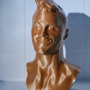 May include: A bronze-colored bust of a smiling person with short, styled hair. The sculpture is a detailed representation of a human face and upper torso, with visible facial features and neck.