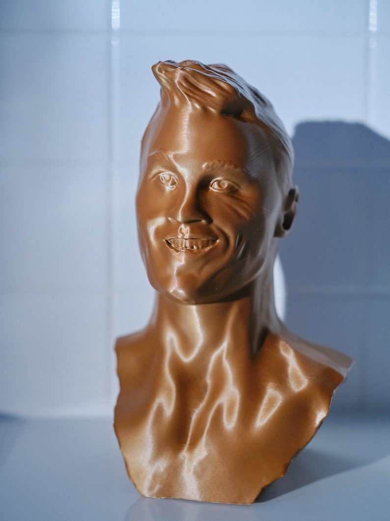 May include: A bronze-colored bust of a smiling person with short, styled hair. The sculpture is a detailed representation of a human face and upper torso, with visible facial features and neck.