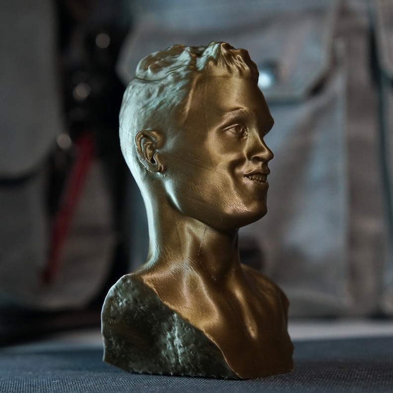 May include: A bronze-colored bust of a person with short hair and a smiling expression. The sculpture is detailed, showing the contours of the face, neck, and shoulders. The bust is likely made of a metallic material.