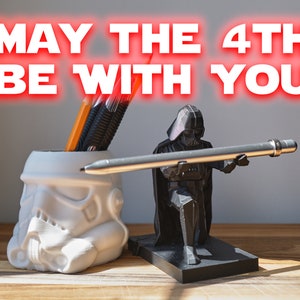 May include: A white Stormtrooper helmet pencil holder with three pens inside, next to a black Darth Vader figurine holding a silver pen. The background is a light grey wall with the text "MAY THE 4TH BE WITH YOU" in red.
