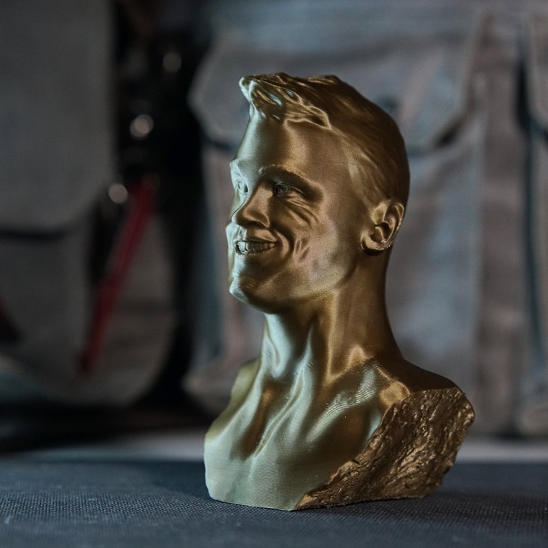 May include: A gold-colored bust of a smiling person, showcasing the head and upper torso. The sculpture has detailed facial features and a textured surface, with the hair styled upwards. The bust is set against a blurred background.