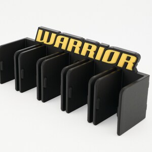 Puede incluir: Organizador de pl&aacute;stico negro con ranuras para guardar objetos. El organizador tiene la palabra "WARRIOR" impresa en amarillo en la parte superior.