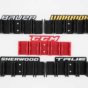 Puede incluir: Un conjunto de cinco soportes para palos de hockey, cada uno con un nombre de marca diferente. Los soportes son negros con letras blancas, amarillas y rojas. Las marcas son Bauer, Warrior, CCM, Sherwood y True.
