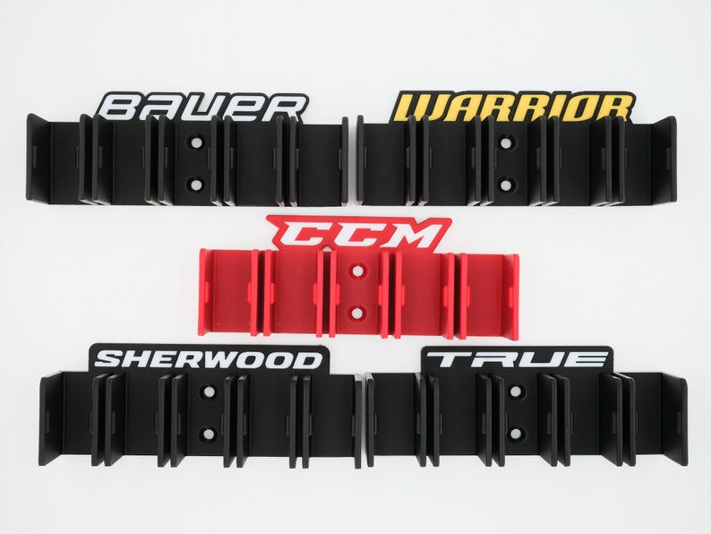 Puede incluir: Un conjunto de cinco soportes para palos de hockey, cada uno con un nombre de marca diferente. Los soportes son negros con letras blancas, amarillas y rojas. Las marcas son Bauer, Warrior, CCM, Sherwood y True.