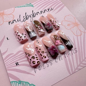 Pu&ograve; includere: Un set di dieci unghie press-on con un tema tropicale. Le unghie sono rosa con vari disegni tra cui fiori, stampa animalier e personaggi dei cartoni animati. Le unghie sono esposte su una carta rosa con il testo "nailsbybanani" e "press on nails".