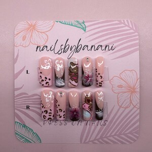 Pu&ograve; includere: Un set di 10 unghie finte con base rosa e vari design, tra cui un personaggio di Hello Kitty, fiori e stampa leopardata. Le unghie sono esposte su una carta rosa con il testo "nailsbybanani" e "PRESS ON NAILS".