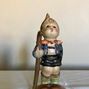 Pode incluir: Uma figurinha de porcelana de um menino vestindo uma jaqueta azul, uma echarpe vermelha e branca e calções pretos. Ele está segurando um pau marrom e tem um penteado castanho claro para cima. A figurinha está em pé sobre uma base branca.