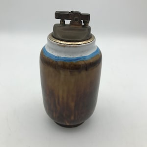 May include: A brown ceramic table lighter with a blue and white band around the top. The lighter has a metal top with a flip-top lid.
