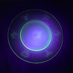 Può includere: Un piatto di vetro verde con un motivo a grappolo d'uva. Il piatto è illuminato da una luce nera, che lo fa brillare.