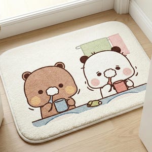 May include: A rectangular, off-white bath mat featuring a cartoon illustration of two bears drinking from mugs. The bears are brown and white, with pink cheeks. A blue line represents water, and two towels hang above. The mat is placed on a wooden floor.
