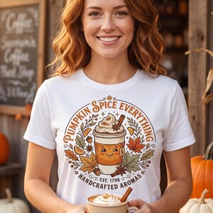 Könnte beinhalten: Ein weißes T-Shirt mit einer Grafik einer Kaffeetasse mit lächelndem Gesicht, umgeben von Herbstblättern, Zimtstangen und dem Text "Pumpkin Spice Everything" und "Handcrafted Aromas". Das T-Shirt wird von einer Person gehalten.