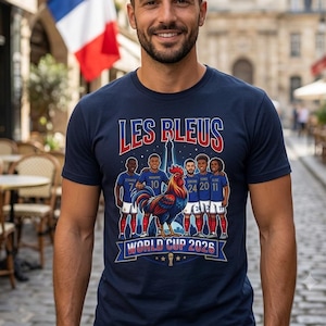 May include: A navy blue short-sleeve t-shirt featuring a graphic print with the text LES BLEUS and WORLD CUP 2026. The design includes five soccer players in blue uniforms, a large colorful rooster, and the Eiffel Tower in the background.