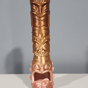 May include: A gold and rose gold tiki statue with a carved face and a hollowed out area in the bottom. The statue is made of a 3D printed material.