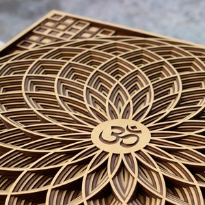 May include: A wooden wall art piece with a layered design of a flower with an Om symbol in the center. The flower is made of multiple layers of wood, creating a 3D effect.