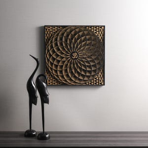 May include: A black square wooden wall art piece with a geometric design in a light wood color. The design features a circular pattern with an Om symbol in the center. Two black bird sculptures are in the foreground.