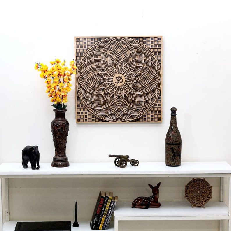 May include: A wooden wall art piece with a mandala design featuring the Om symbol in the center. The mandala is surrounded by a square frame with a checkered pattern. The art piece is displayed on a white wall above a shelf with various decorative items.