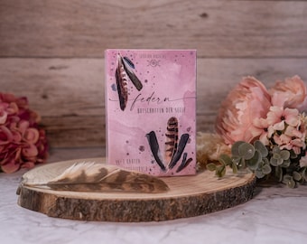 May include: A pink box with the text "Federn Botschaften der Seele" and a watercolor illustration of feathers. The box is on a wooden surface with a feather next to it.