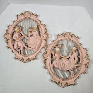 May include: Two vintage wall plaques featuring relief sculptures of couples in period clothing. The oval frames are light pink with gold accents and a black mesh background. The sculptures depict figures in pink attire, evoking a romantic, classical theme.