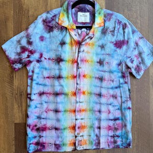 May include: A short-sleeved button-down shirt with a vibrant tie-dye pattern in shades of blue, purple, yellow, and orange. The shirt has a collar and is hanging on a black hanger. The brand label "HOLLISTER" is visible.