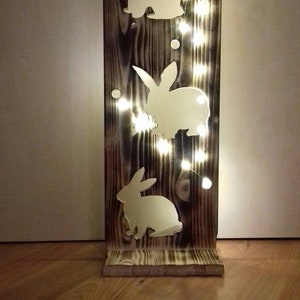 Könnte beinhalten: Eine Holzlampe mit drei ausgeschnittenen Hasensilhouetten. Die Lampe ist mit weißen Lichterketten beleuchtet.