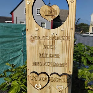 Könnte beinhalten: Holzhochzeitsschild mit kreisförmigem Ausschnitt und herzförmigen Details. Das Schild enthält die Namen "LYDIA" und "ACHIM", das Datum "05.JULI.2025" und den Text "DER SCHONSTE WEG IST DER GEMEINSAME". Ein Herz mit dem Namen "LEO" hängt im Kreis.