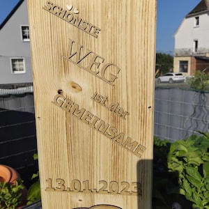 Könnte beinhalten: Holzschild mit lasergeschnittenem deutschen Text, darunter "DER SCHÖNSTE WEG ist der GEMEINSAME" und das Datum 13.01.2023. Mit einem Design aus zwei ineinandergreifenden Ringen. Das Schild ist aus hellem Holz gefertigt.