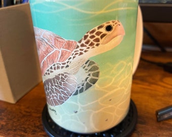 May include: A white ceramic mug with a green, blue, and brown sea turtle design. The turtle is swimming in a turquoise ocean with white waves. The mug is sitting on a black coaster.