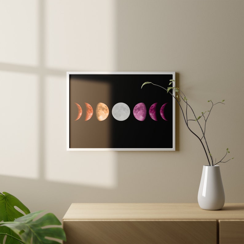 May include: A framed print of a black background with a series of six moons in different phases, colored in a lesbian pride flag color scheme. The moons are in a row from left to right, starting with a crescent moon in red, then a half moon in orange, a full moon in white, a half moon in pink, and a crescent moon in purple.