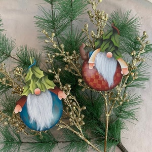 Peut inclure: Deux décorations de gnome peintes à la main avec des chapeaux verts et des barbes blanches. Les gnomes sont décorés de motifs à carreaux rouges et blancs et sont attachés à une brindille dorée.